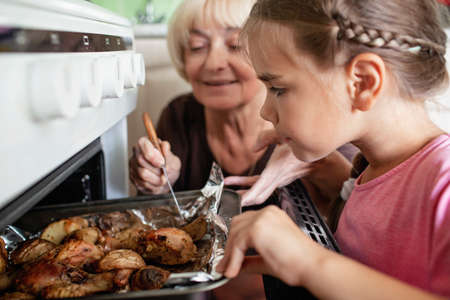 Cooking for family dinner. Excited caring grandmother and small girl taking pan with baked roast chicken and potato out of oven, together in the kitchen, home lifestyleの写真素材