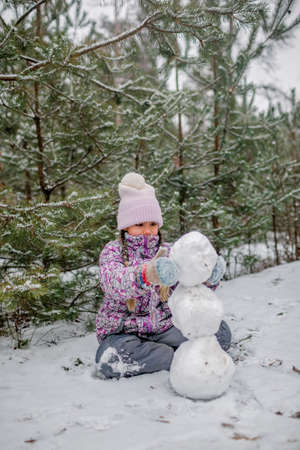 Pretty girl in winter clothes makes a snowman from the first long-awaited snow during family walking in forest on snowing day, outdoor lifestyle, winter wonderlandの写真素材