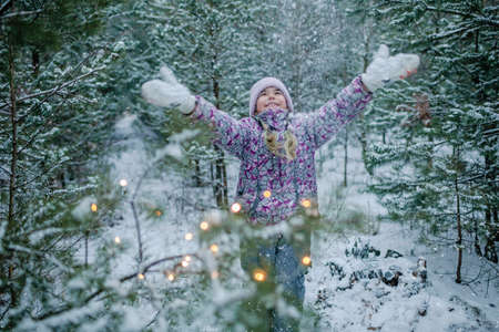 Pretty girl in warm clothes, with frozen mittens dances among the first long-awaited snow during family walking in forest on snowing day, outdoor lifestyle, winter wonderlandの写真素材