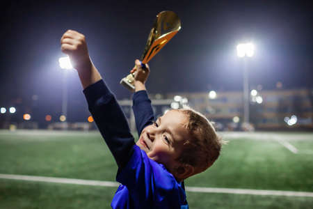 Young soccer player in blue jersey with ten number raising a trophy after the winning goal in the football competition, illuminated stadium, sport, winner and success. Dreams come trueの写真素材