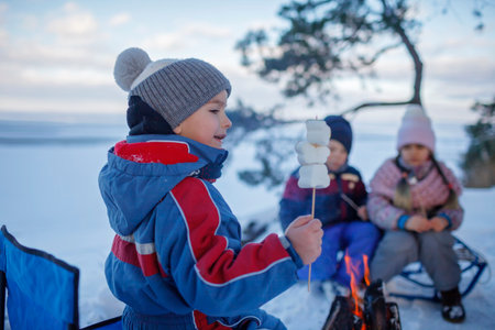 Happy friends sit around campfire on the shore of frozen lake in forest and fry marshmallows during a winter trip at weekend. Family winter picnicの写真素材