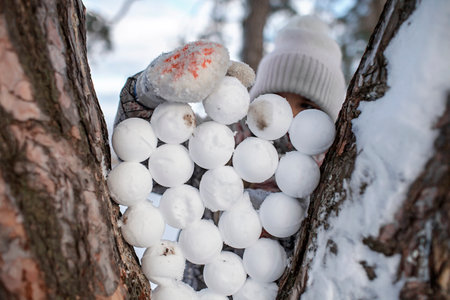 Pretty girl fills the space between the branches of tree with snowballs, fun activity in snow-covered winter forest with family and friends. Seasonal walk, lifestyleの写真素材