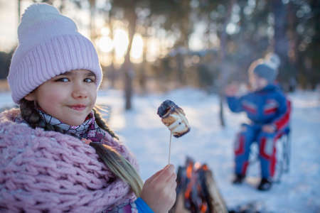 Portrait of happy girl sitting by the fire in snow-covered forest with fried marshmallows, winter trip at weekend. Family winter picnic, active lifestyleの写真素材