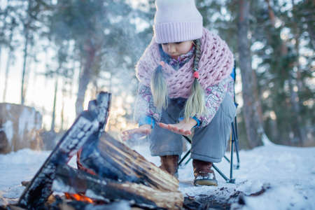 The girl in winter clothes warms herself up by the fire in snow-covered forest, winter trip at weekend. Family winter picnic, active lifestyle, focus on handの写真素材