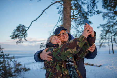 Middle aged man and woman in military jacket with camouflage pattern look at smartphone camera to make selfie, reunion of couple on the shore of beautiful frozen lake in winterの写真素材