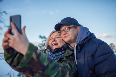 Reunion of couple on the shore of beautiful frozen lake in winter. Middle aged man and woman in military jacket with camouflage pattern look at smartphone camera to make selfieの写真素材