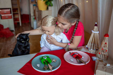 Pretty girl with doll at the table with Christmas setting, cutlery and DIY napkin ring holders like Santa clothes made with toilet paper rolls, festive dinner. Ecological and sustainable celebrationの写真素材