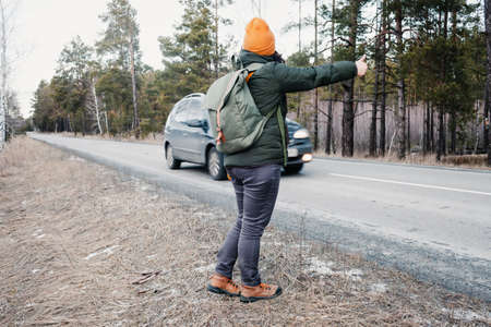 Middle-aged active woman with backpack hiking in the road and gesture trying to stop a car, forest background, winter travel concept, healthy lifestyle, hitchhiking solo tourismの写真素材