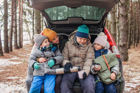Winter family road trip. Happy parents with kids drink coffee in the trunk of car during walking in snowy pine forest, travel together in any season, active lifestyle, authentic peopleの写真素材