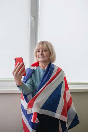Happy senior woman holding British flag makes selfie to share participation in Platinum Jubilee of...