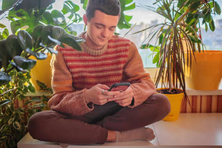 Young man in a cozy sweater sitting on the table and chatting on smartphone in his favorite place...