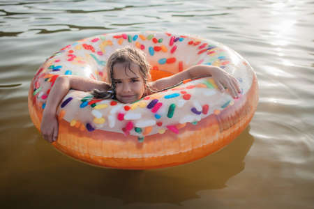 A smiling tween girl swims with a big donut shaped inflatable ring on a lake on a hot summer day, happy summertime, local vacations, cottagecore, non-urban landscapeの写真素材