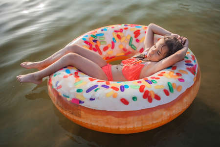 A smiling tween girl relaxing on a big donut shaped inflatable ring on a lake on a hot summer day, happy summertime in countryside, local vacations, cottagecoreの写真素材