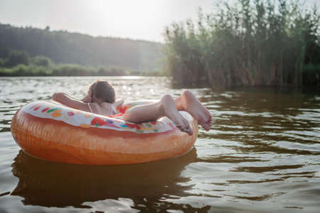 A smiling tween girl relaxing on a big donut shaped inflatable ring on a lake on a hot summer day, happy summertime in countryside, local vacations, cottagecoreの写真素材