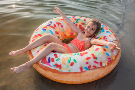 A smiling tween girl having fun on a big donut shaped inflatable ring on a lake on a hot summer day, happy summertime in countryside, local vacations, cottagecoreの写真素材