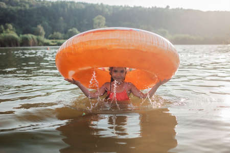 A smiling tween girl having fun on a big donut shaped inflatable ring on a lake on a hot summer day, happy summertime in countryside, local vacations, cottagecoreの写真素材