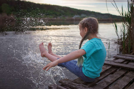 Happy little girl sitting on pontoon bridge on the bank of lake, splashing water and enjoying warm sunny evening, childhood in village in summer, countrysideの写真素材