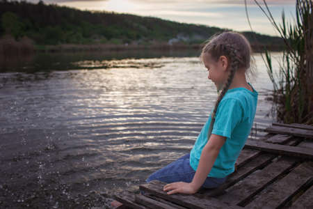 Happy little girl sitting on pontoon bridge on the bank of lake and enjoying warm sunny evening, childhood in village in summer, countrysideの写真素材