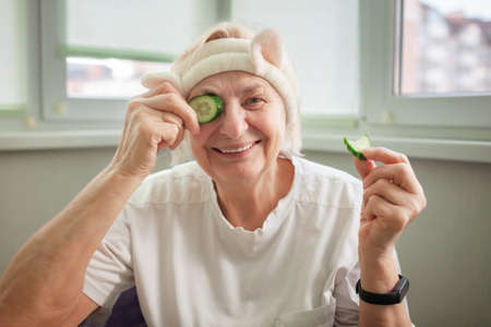 Cheerful mature woman covers eyes with green cucumber and enjoys life at home, wellness and beauty care at any age, body positivity, self-esteem and ageism, lifestyleの写真素材