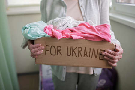 Senior Ukrainian woman looking at a cardboard box with clothes for Ukrainian refugees fleeing from Russian invasion. World terrorism, war and current historyの写真素材