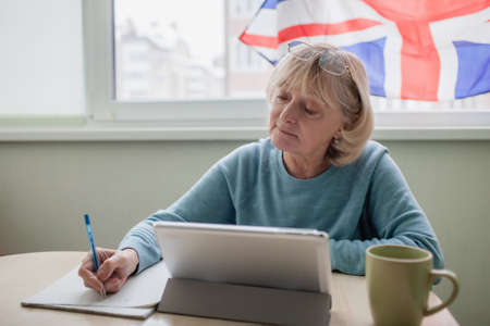 Senior woman writes new English words during distant learning with laptop, language classes for Ukrainian refugee for better adaptation abroad, British flag on the window, social distance educationの写真素材