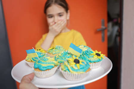 Excited Ukrainian girl for whom the neighbors brought for brunch cupcakes with cream in yellow and blue colors and decorated with the flag of Ukraine. Support for Ukrainian refugees around the worldの写真素材