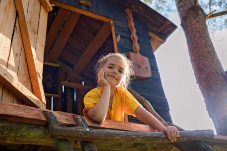 Happy pretty girl looking down from the beautiful creative handmade treehouse in backyard, summer activity, cottagecore, happy summertime in countryside, ecological outdoor playground, lower angleの写真素材