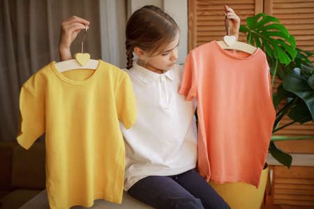 Pretty girl chooses T-shirt among clothes at home, she holds light colored yellow and pink plain ones. Sale, donation, fashion, wardrobe renovation. Mockup friendly. T-shirts ready to have designの写真素材