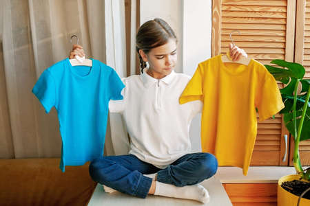 Pretty girl chooses T-shirt among clothes at home, she holds light colored yellow and blue plain ones. Sale, donation, fashion, wardrobe renovation. Mockup friendly. T-shirts ready to have designの写真素材