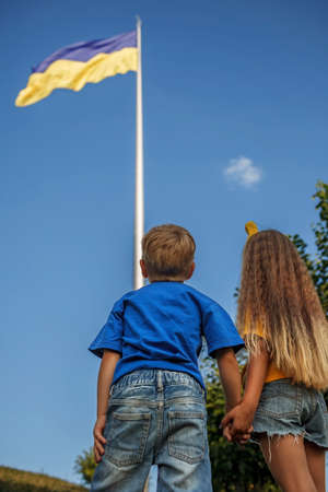 Small patriots, girl in yellow and boy in blue, looking at the biggest national flag of Ukraine. Brave Ukrainian nation defends its independence and believes in victory. Clean blue sky backgroundの写真素材