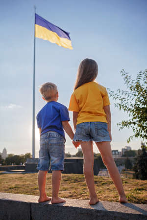 Small patriots, girl in yellow and boy in blue, looking at the biggest national flag of Ukraine. Brave Ukrainian nation defends its independence and believes in victory. Clean blue sky backgroundの写真素材