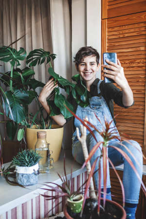 Happy young girl makes photo with smartphone to share in social media, home plants on the balcony, plant influencer, home gardening, green decor and environment at home, generation Z trendの写真素材
