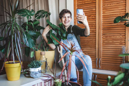 Happy young girl makes photo with smartphone to share in social media, home plants on the balcony, plant influencer, home gardening, green decor and environment at home, generation Z trendの写真素材