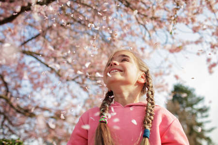 A happy girl in a pink hoodie is throwing sakura petals like those that its snowing in spring. She...