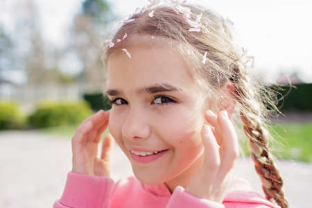Emotional portrait of happy girl in a pink hoodie with sakura petals on her head and eyelashes that fell from trees when the wind blew. She rejoices in the coming of springtime. Spring vibes.の写真素材