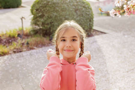 A happy girl in a pink hoodie is ready to throw sakura petals into the camera. She rejoices in the coming of spring. Spring vibes. Emotional lifestyle portraitの写真素材