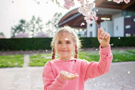 A happy girl in a pink hoodie is throwing sakura petals like those that its snowing in spring. She rejoices in the coming of spring. Spring vibes. Emotional lifestyle portrait. Selective focusの写真素材