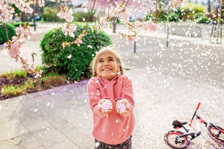A happy girl in a pink hoodie is throwing sakura petals like those that its snowing in spring. She rejoices in the coming of spring. Spring vibes. Emotional lifestyle portrait. Selective focusの写真素材