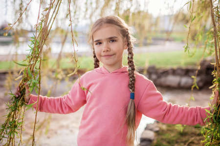 Preteen girl in pink hoodie able to have design walks at the bank of lake among green willows in early spring, relax and freedom. Happy kid enjoying spring and sunny day. Outdoor portrait.の写真素材