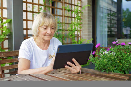 Senior woman reading news with tablet on the cozy balcony at home. Active retirement, baby boomer generations. Senior people with digital technologies. Terrace life in spring and summerの写真素材