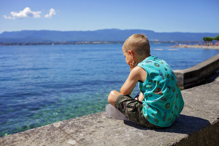 A boy sits on a parapet on the quay of Geneva Lake and looks at the water and mountains, sunny summer day, vacation and travel, lifestyle, view from backの写真素材