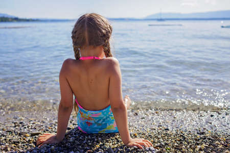 Cute girl has a rest and sits on stones on pebble beach at Geneva Lake over blue mountains in sun light, daydreaming and happiness, summer vacations, travel and summertime, lifestyle, back viewの写真素材