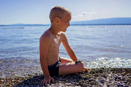 Happy boy has fun and plays with stones on pebble beach at Geneva Lake over blue mountains in sun light, lots of fun and happiness, summer vacations, travel and summertime, lifestyleの写真素材