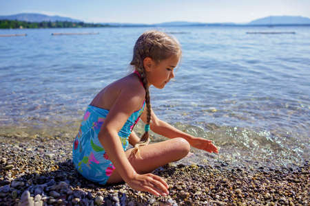 Happy girl has fun and plays with stones on pebble beach at Geneva Lake over blue mountains in sun light, lots of fun and happiness, summer vacations, travel and summertime, lifestyleの写真素材