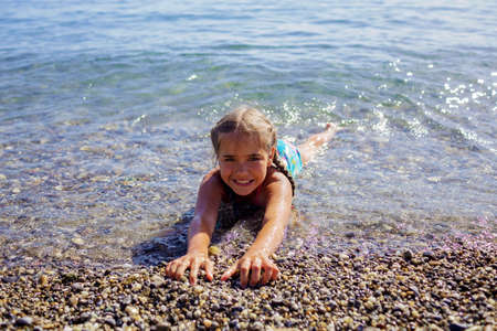 A happy girl lies in the water on the pebbly beach of Geneva Lake over mountains and a blue sky, lots of fun and happiness, summer vacations, travel and summertime, lifestyleの写真素材
