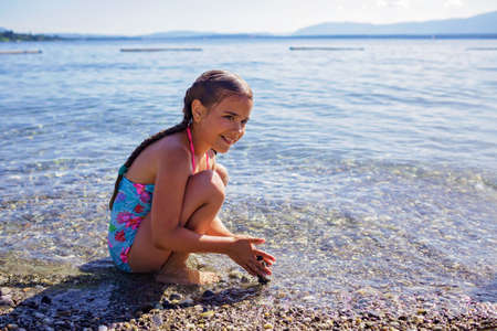 Happy girl has fun and plays with stones on pebble beach at Geneva Lake over blue mountains in sun light, lots of fun and happiness, summer vacations, travel and summertime, lifestyleの写真素材