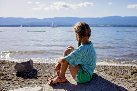 A girl sits on the stone on the shore of Geneva Lake and looks at the water and mountains, sunny summer day, vacation and travel, lifestyle, view from backの写真素材