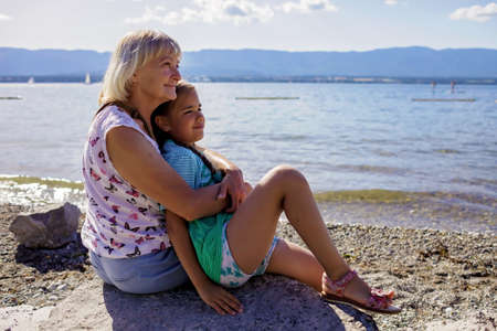 A senior woman with her grandkid sits on the stone on the shore of Geneva Lake and looks at the water and mountains, sunny summer day, family vacation and travel, lifestyleの写真素材