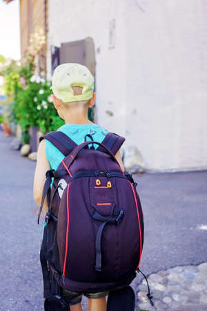 A small tourist with backpack walks along the street in typical European small village with old stone squares and courtyard with flowers, summer vacations, family travel, view from backの写真素材