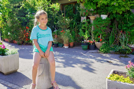 Girl sits and waits for a bus in typical European small village with old stone squares and courtyard with flowers, summer vacations, family travel, beautiful summertimeの写真素材